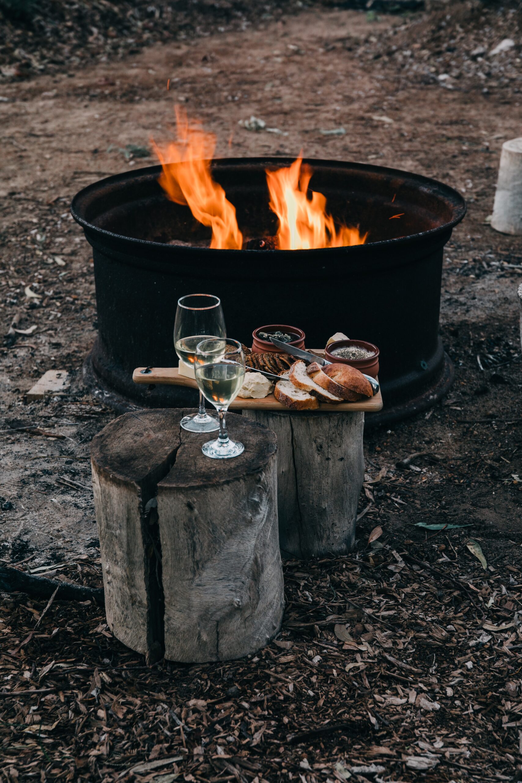 Image represents time off. A bonfire burns near tree stumps. On the stumps are snacks and wine glasses. 